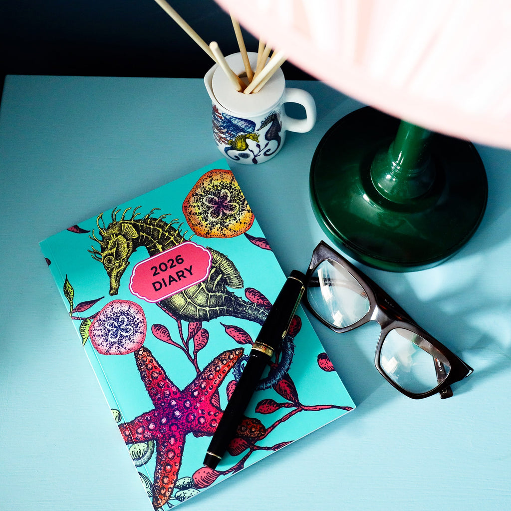 Cream Cornwall 2026 reef diary with seahorse and starfish design, styled with pen, glasses and lamp on blue desk
