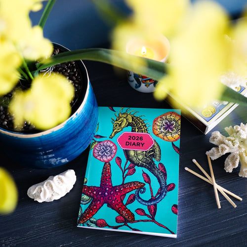 Cream Cornwall 2026 reef diary with colourful seahorse and starfish cover, styled with candle, coral and flowers on a dark table.