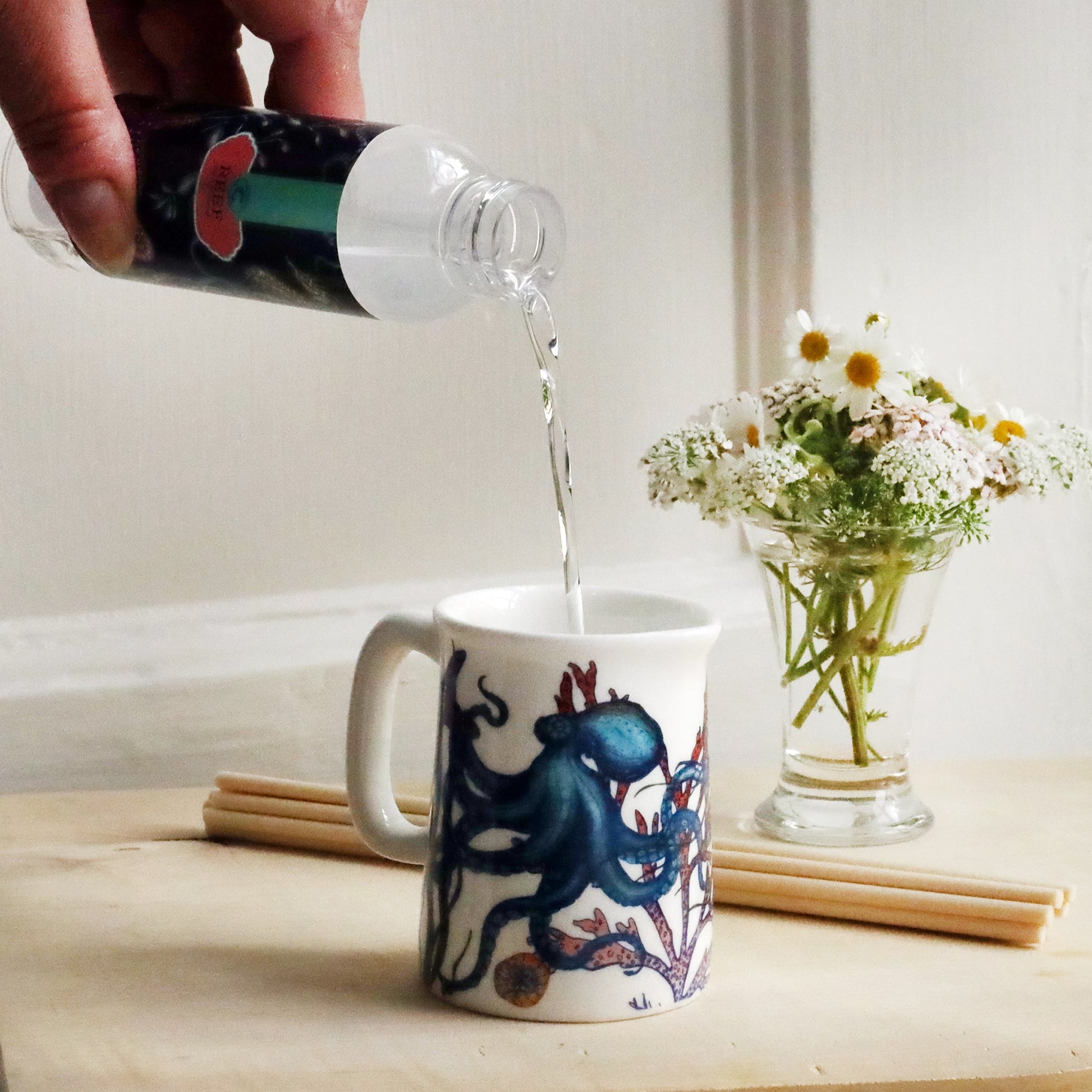 Mini Jug with octopus reef design on with reeds and small posy of flowers behind with a bottle of Reef room diffuser oil being poured into the jug.