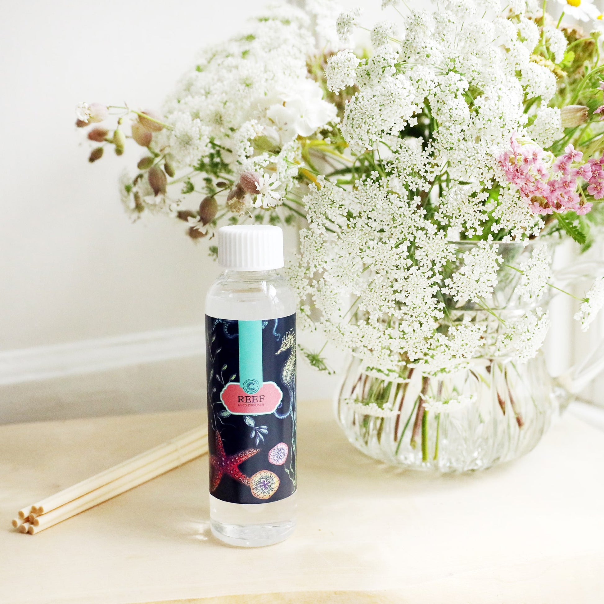 Bottle of Reef room diffuser oil with octopus design on the label, sitting on a wooden block with the diffuser reeds laying jut to the side, and a vase of soft coloured flowers sitting behind.
