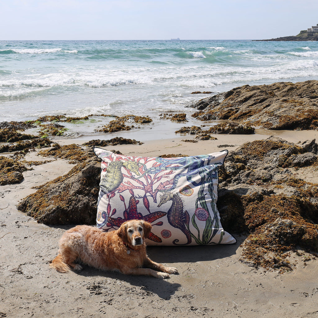 Dog lying on a sandy beach with a decorative pillow featuring ocean-themed design.