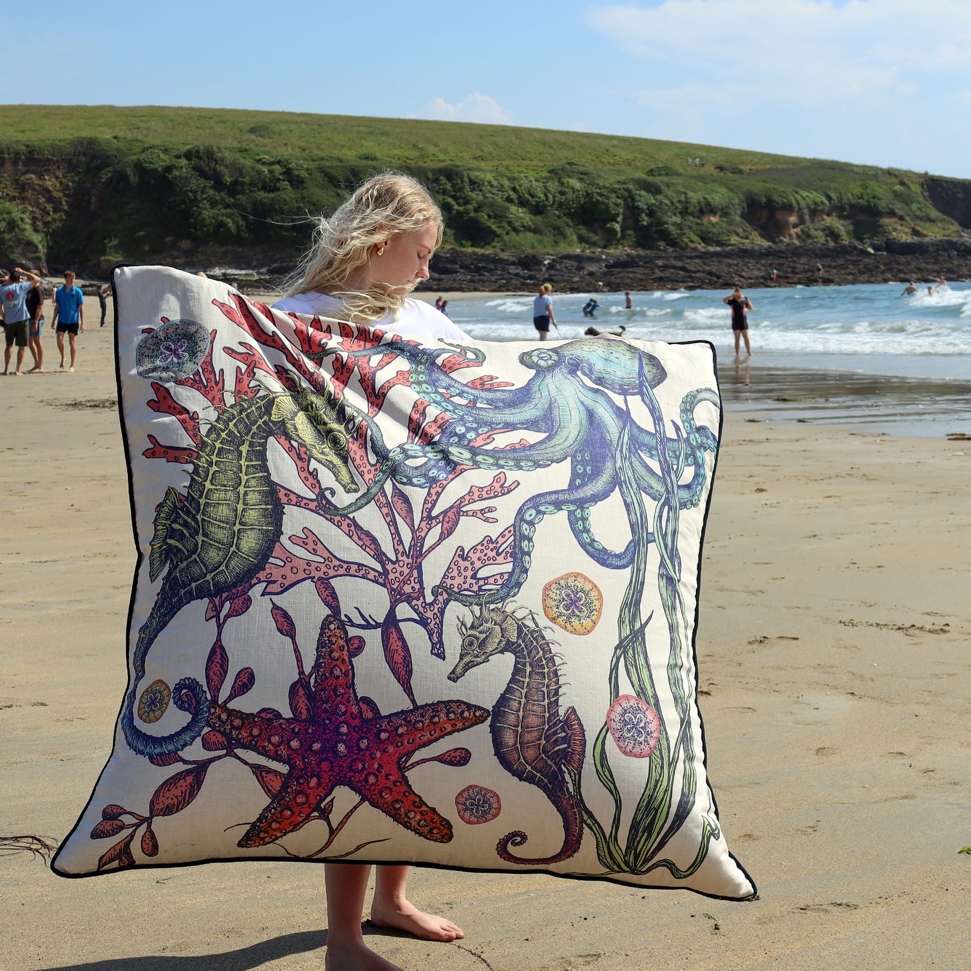 Person holding a large floor cushion with marine life design on a sandy beach.