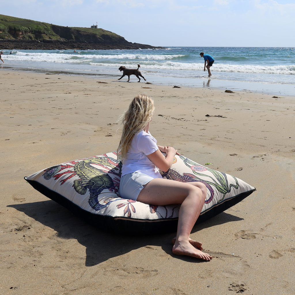 Person sitting on a large decorative pillow on a beach with a dog and person in the background.