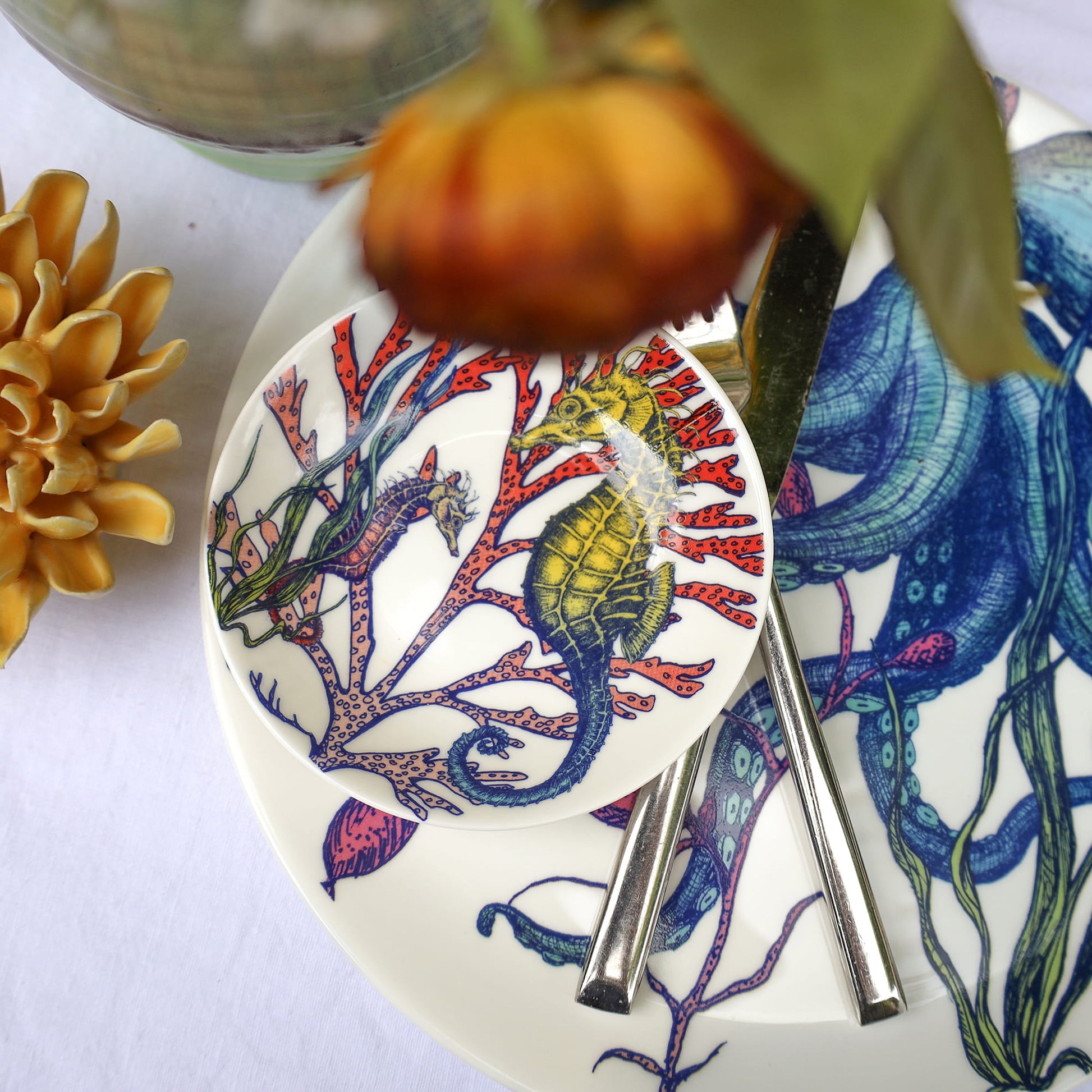 Decorative plate with seahorse design, cutlery, and a small pumpkin on a white surface.