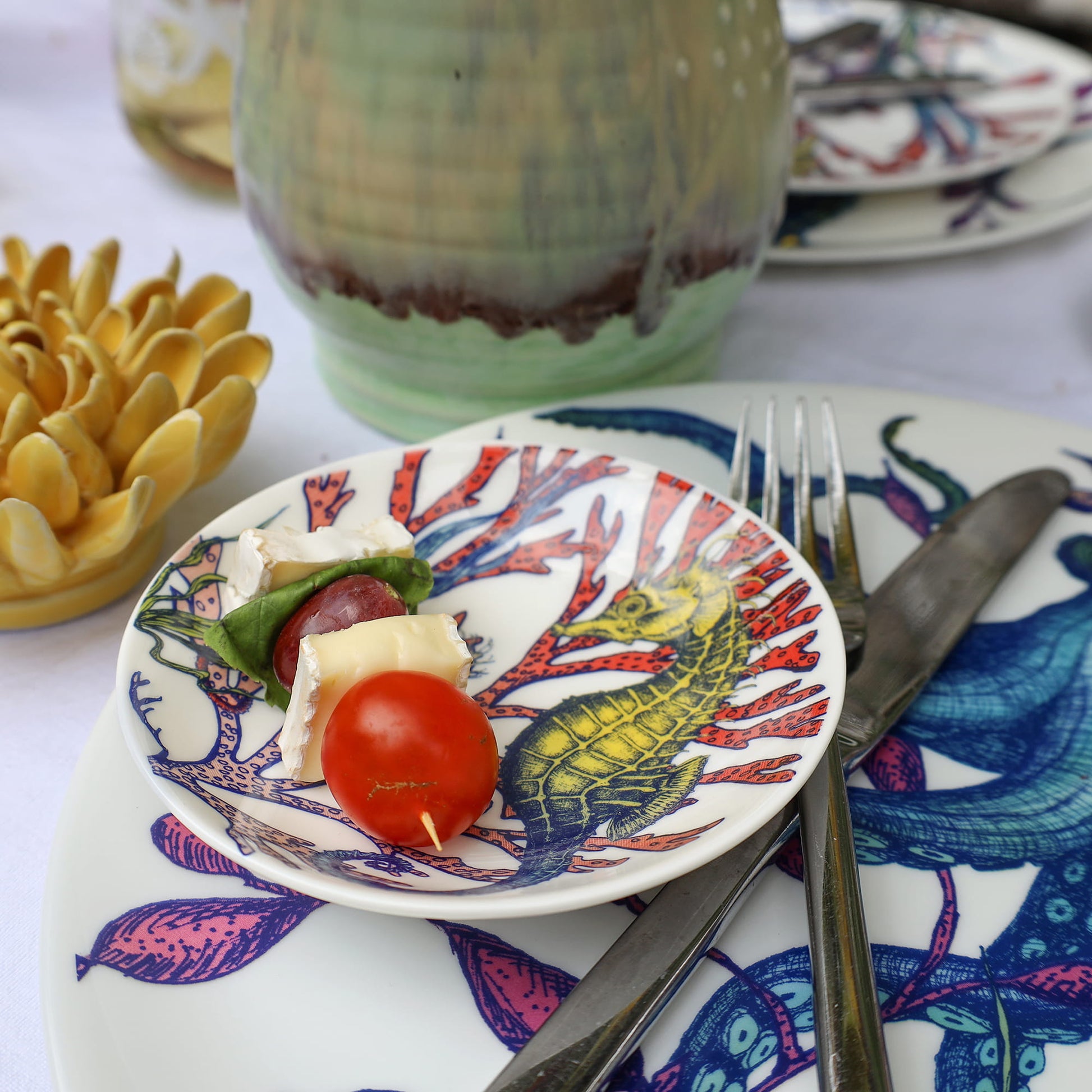 Small plate with food on a decorative plate with a seahorse design, surrounded by cutlery and a ceramic teapot.