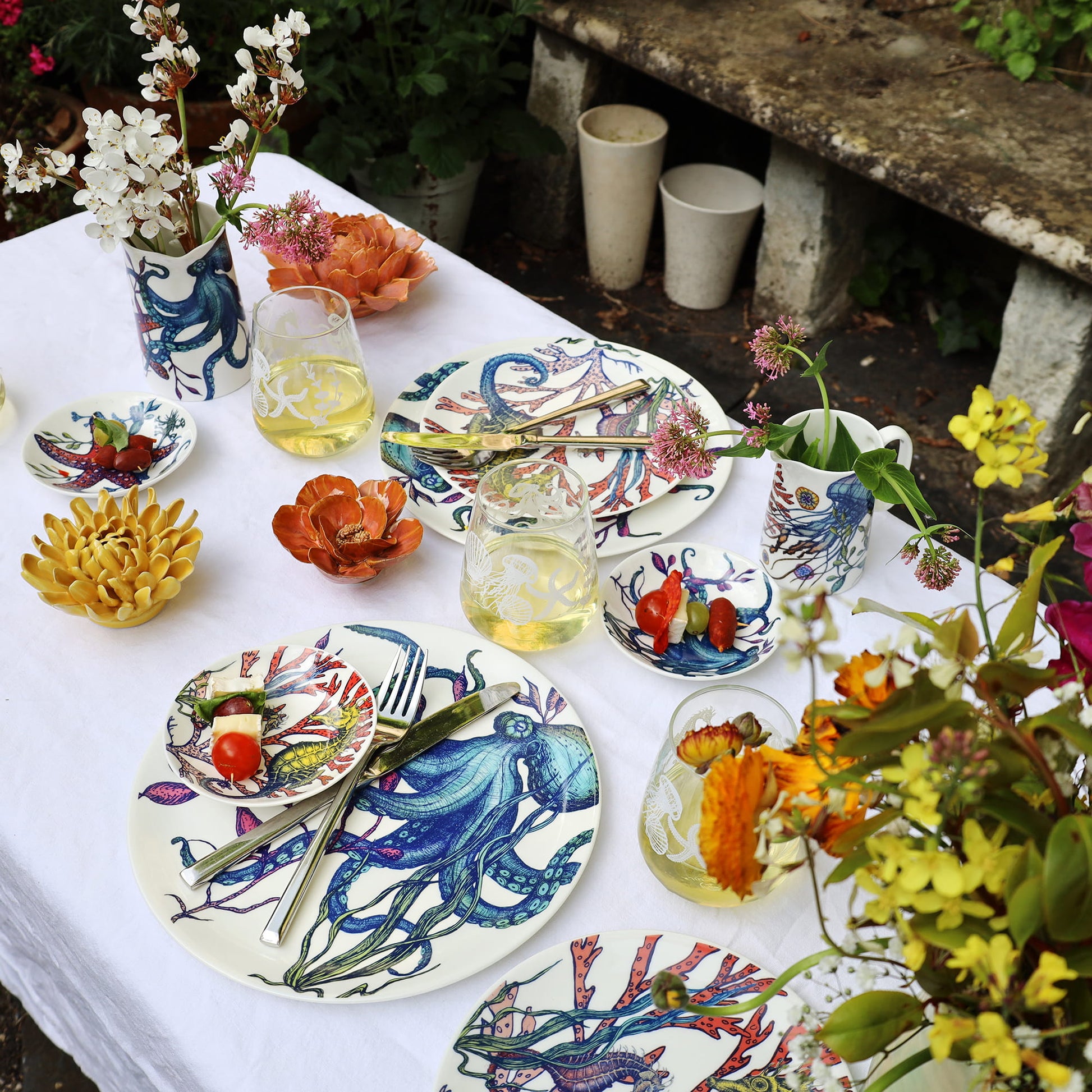 Outdoor table setting with colorful plates, glasses, and floral arrangements.