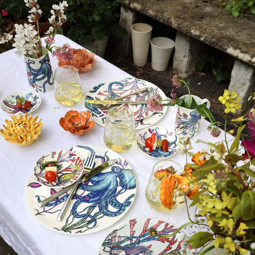 Outdoor table setting with colorful ceramic plates, glasses, and teapot on a white tablecloth.