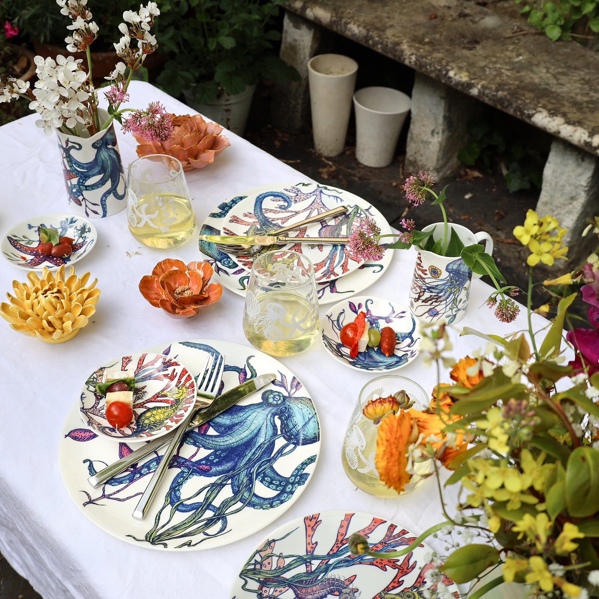 Outdoor table setting with colorful ceramic plates, glasses, and teapot on a white tablecloth.