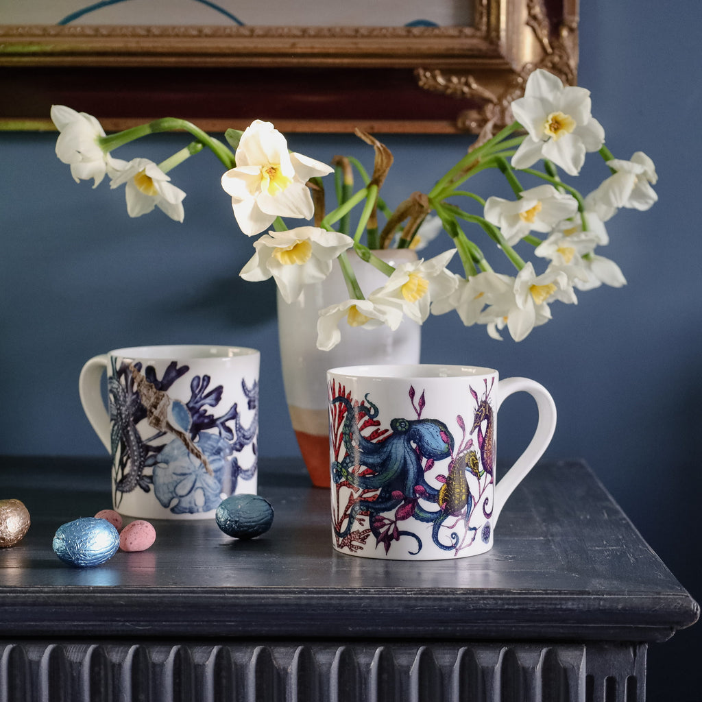 Two decorated mugs on a table with a vase of white flowers and Easter eggs.