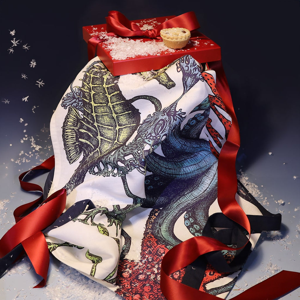 Colourful reef-themed apron featuring illustrations of a seahorse, octopus, and coral, draped beside a red gift box tied with ribbon and topped with faux snow and a mince pie for a festive touch.