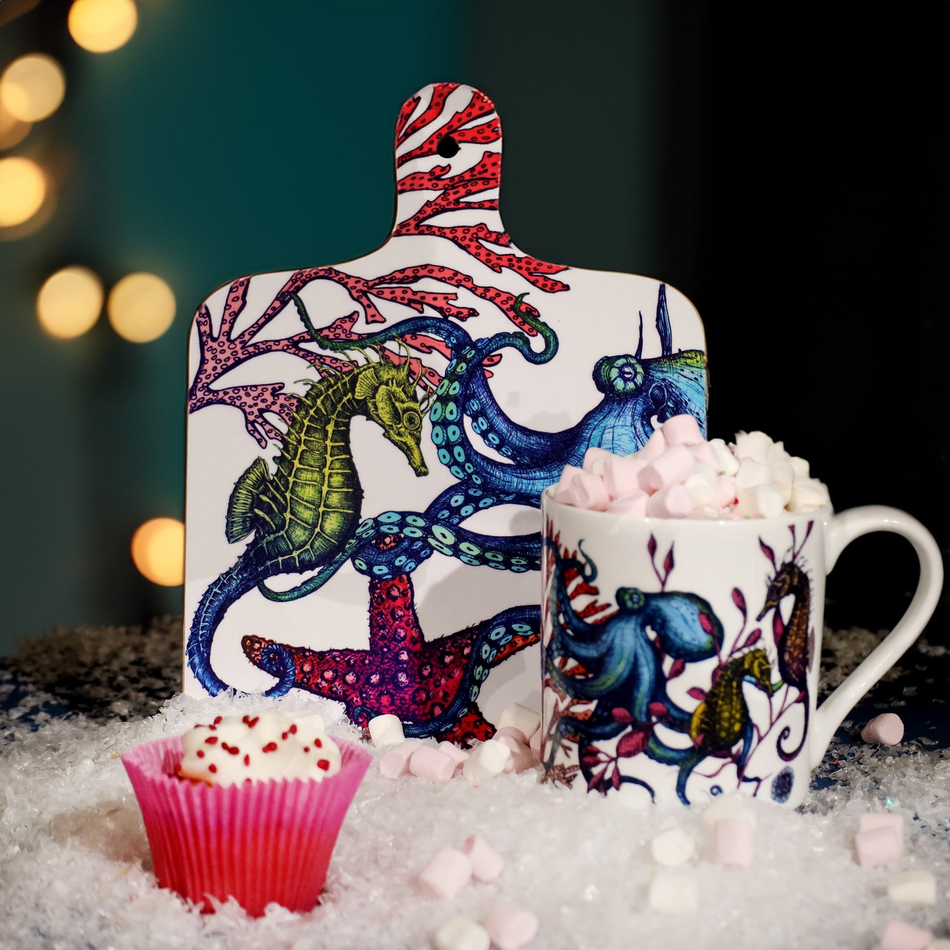 Festive scene featuring a colorful ocean-themed chopping board illustrated with a seahorse, octopus, and starfish, paired with a matching mug filled with hot chocolate and marshmallows, surrounded by faux snow and a pink cupcake with heart sprinkles against a softly lit background.