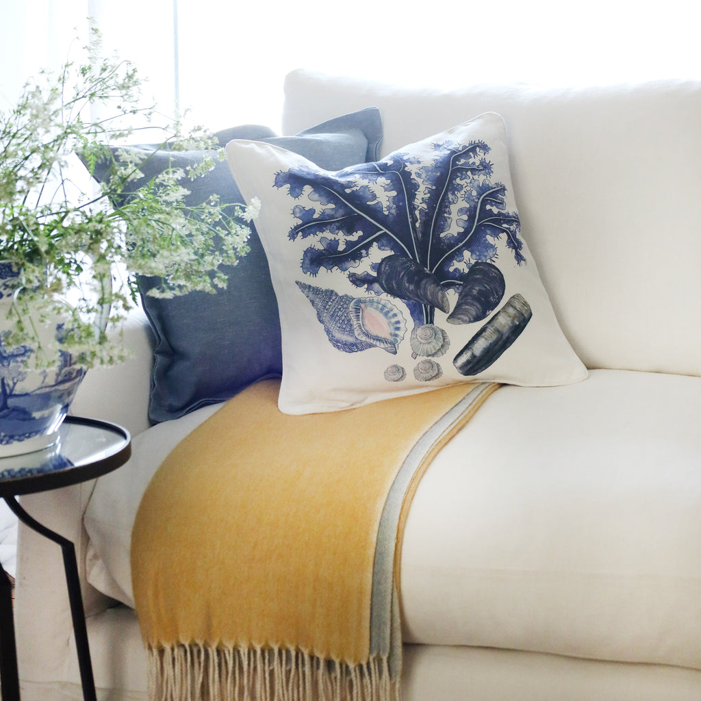 2 cushions an a white sofa with shells and seaweed illustration in tones of blue on with the sun streaming through the window at the back and a large willow pattern jug filled with cow parsley