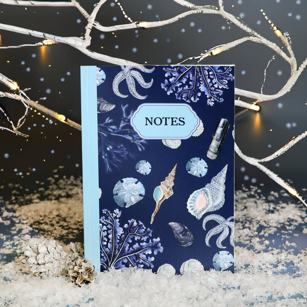 Festive scene featuring a navy blue notebook with seashell and coral illustrations and a light blue spine labeled "NOTES," placed on faux snow with frosted pinecones and surrounded by white branches decorated with twinkling fairy lights.