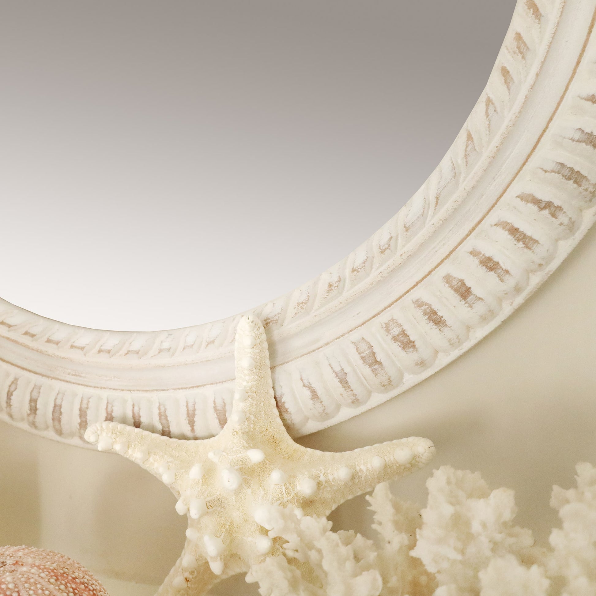 The image is a close-up of a white, textured mirror frame with a rope-like design. In front of the mirror, a decorative starfish is leaning against the frame, surrounded by other coastal-themed decor items, including a piece of white coral and a pink sea urchin. The scene emphasizes natural textures and a soft, beach-inspired color palette.