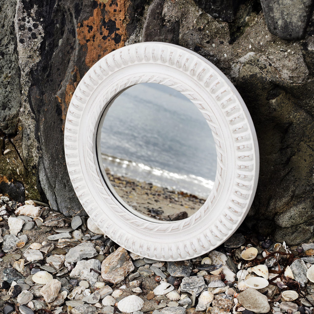 Round whitewashed wooden mirror with rope detail carving propped up against the harbour wall on the beach with the shoreline reflected in it.