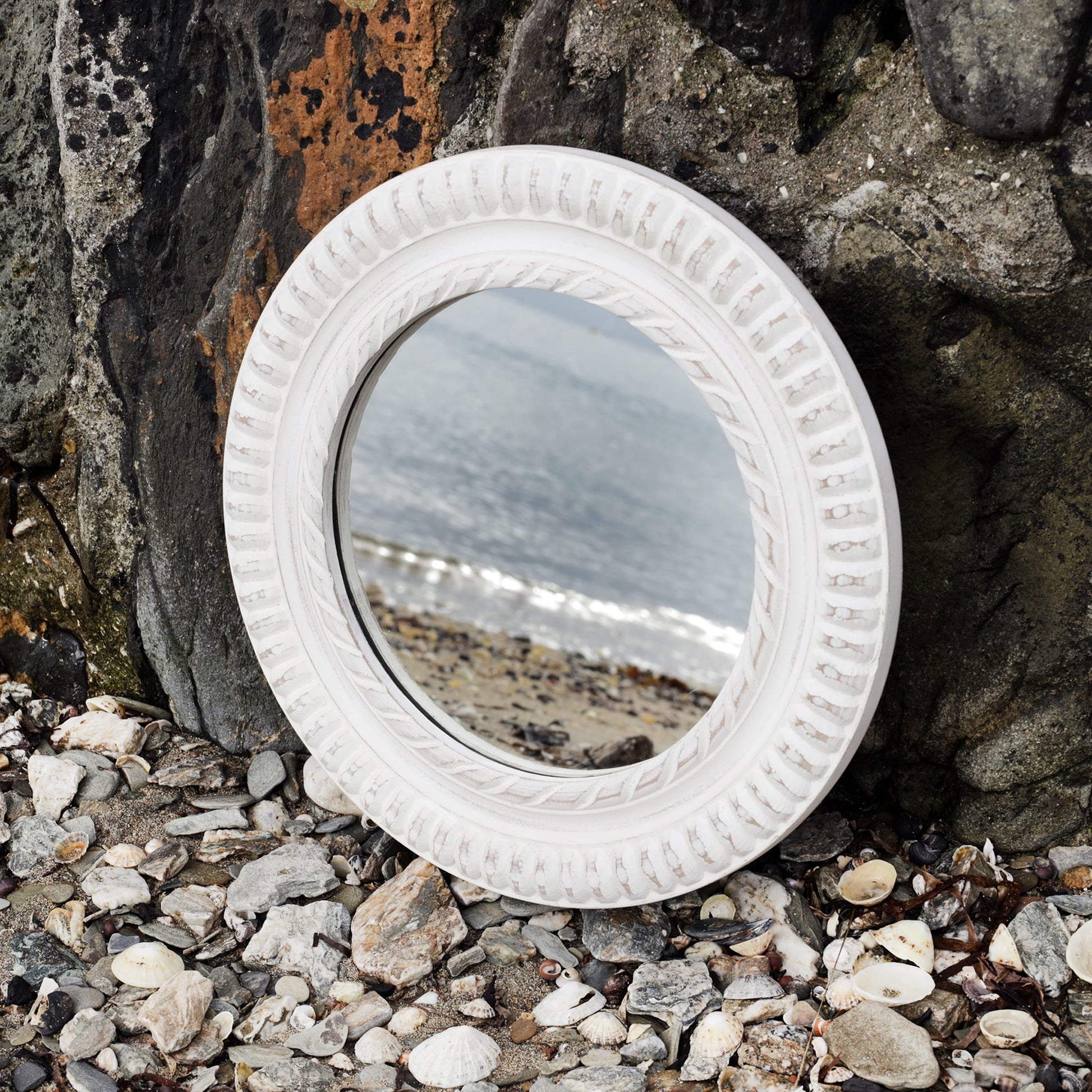 Round whitewashed wooden mirror with rope detail carving propped up against the harbour wall on the beach with the shoreline reflected in it.