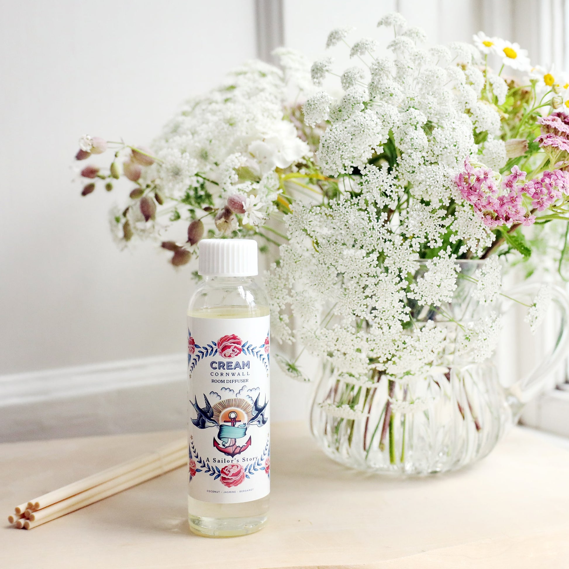 Bottle of Sailor's Story room diffuser oil with octopus design on the label, sitting on a wooden block with the diffuser reeds laying jut to the side, and a vase of soft coloured flowers sitting behind.