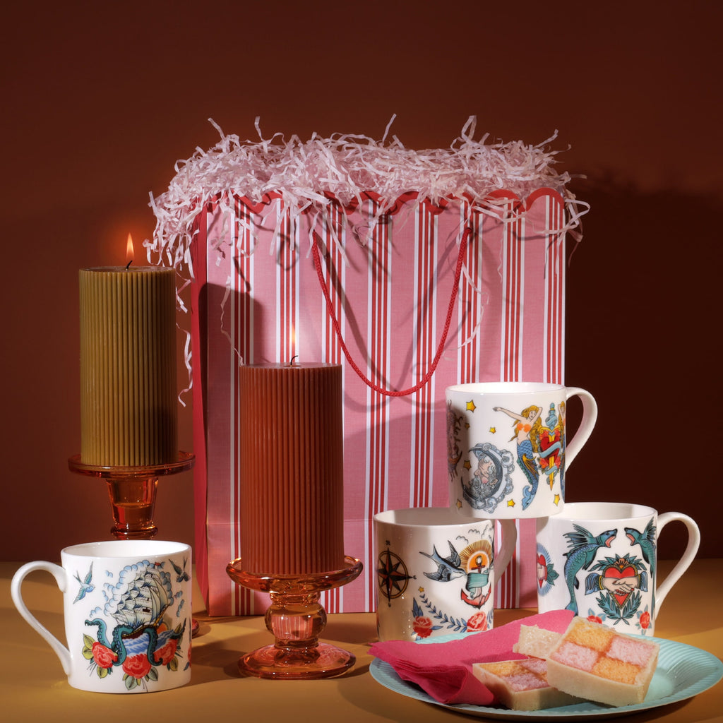 Set of Sailor’s Story mugs with vintage tattoo-inspired designs displayed with candles, a striped gift bag, and plate of sweets.