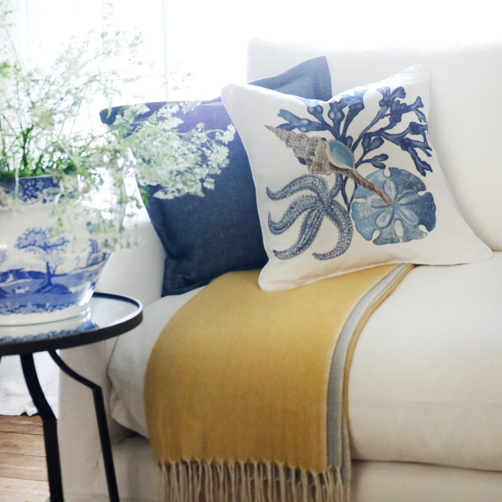 2 cushions an  a white sofa with starfish, shell, seaweed and sand dollar illustration in tones of blue on with the sun streaming through the window at the back and a large willow pattern jug filled with cow parsley