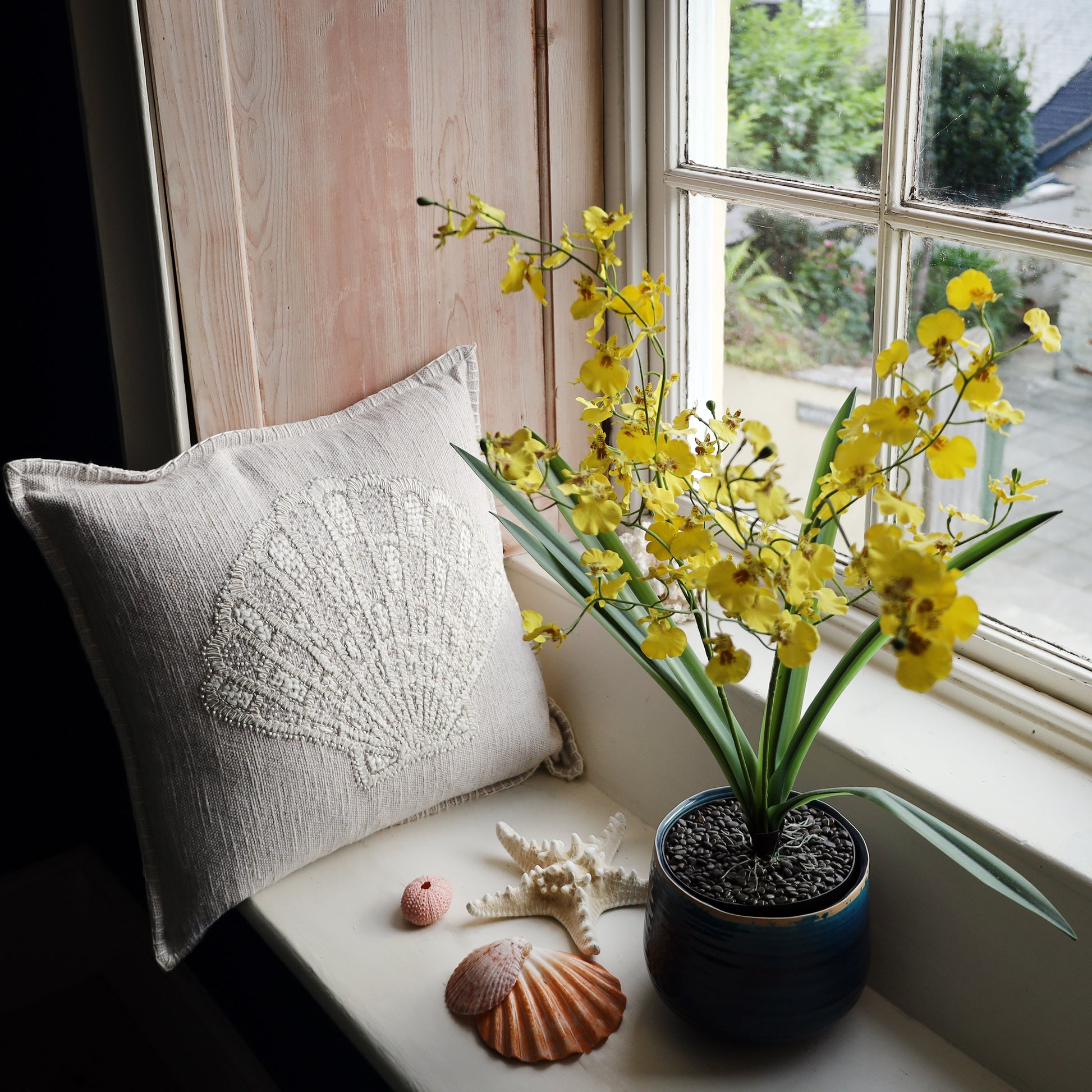 Yellow flowers in a pot on a windowsill with a decorative pillow and shells.