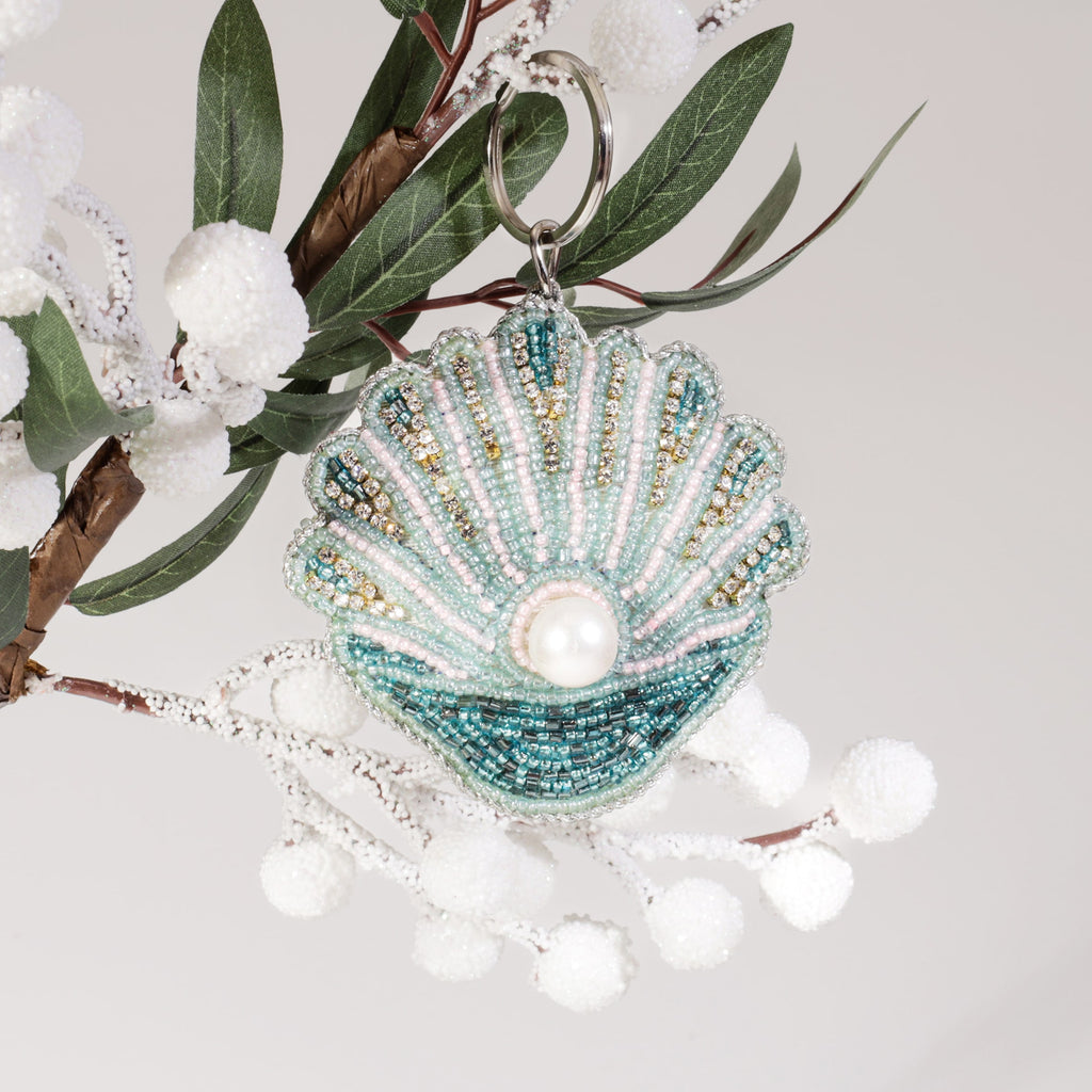 A beaded scallop shell keyring with turquoise, pink, and gold detailing and a central pearl accent, hanging from a branch with white frosted berries and green leaves.
