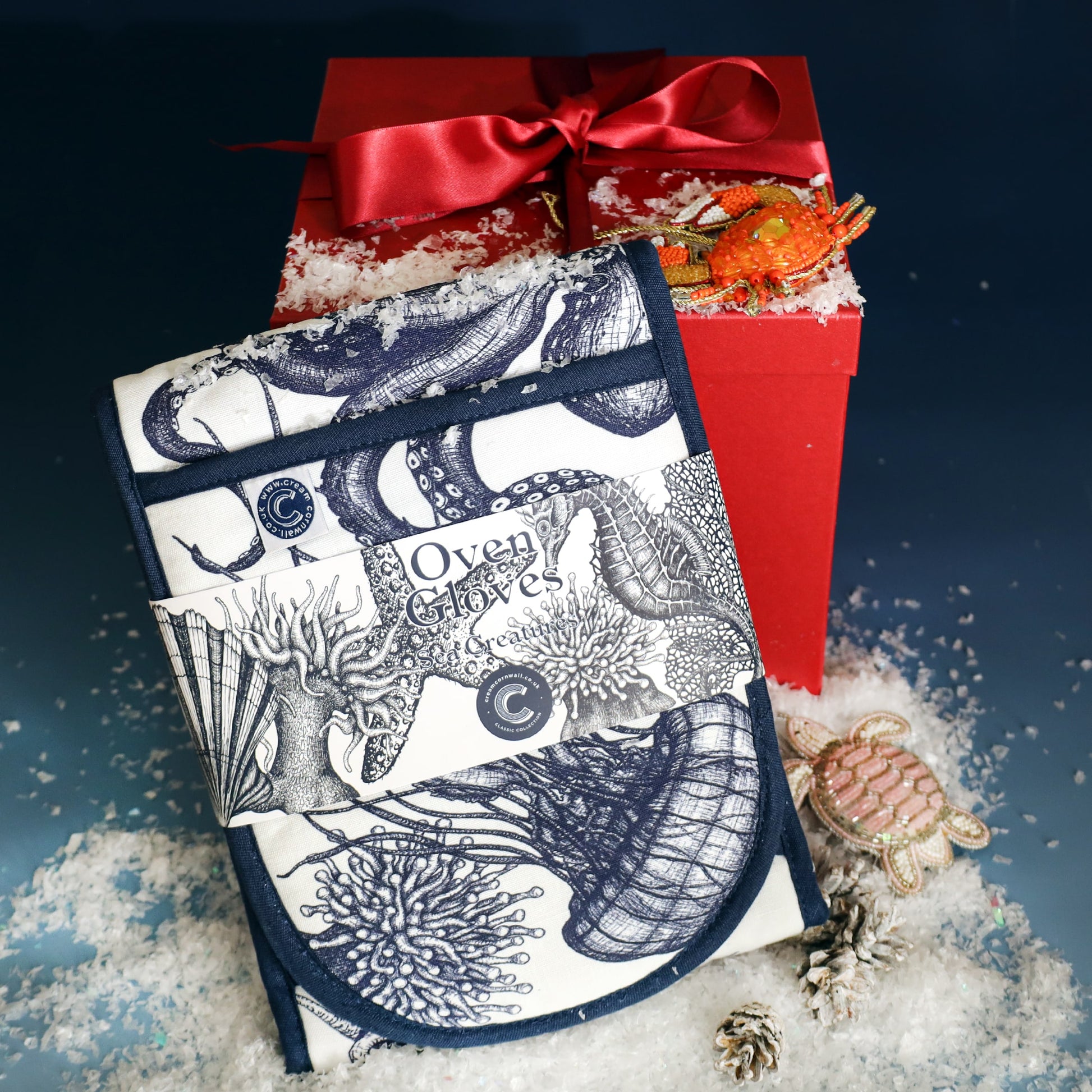Set of Cream Cornwall oven gloves featuring intricate blue sea creature illustrations, displayed with faux snow beside a red gift box tied with a satin ribbon and decorated with beaded crab and turtle ornaments.
