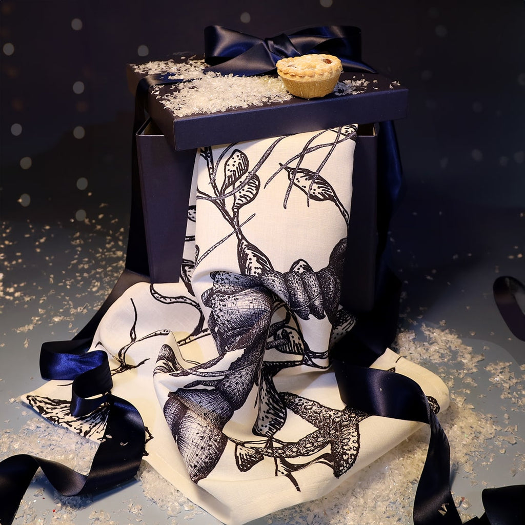 Cream tea towel featuring intricate navy sea creature illustrations, draped over a navy gift box tied with satin ribbon, topped with faux snow and a mince pie for a festive coastal display.