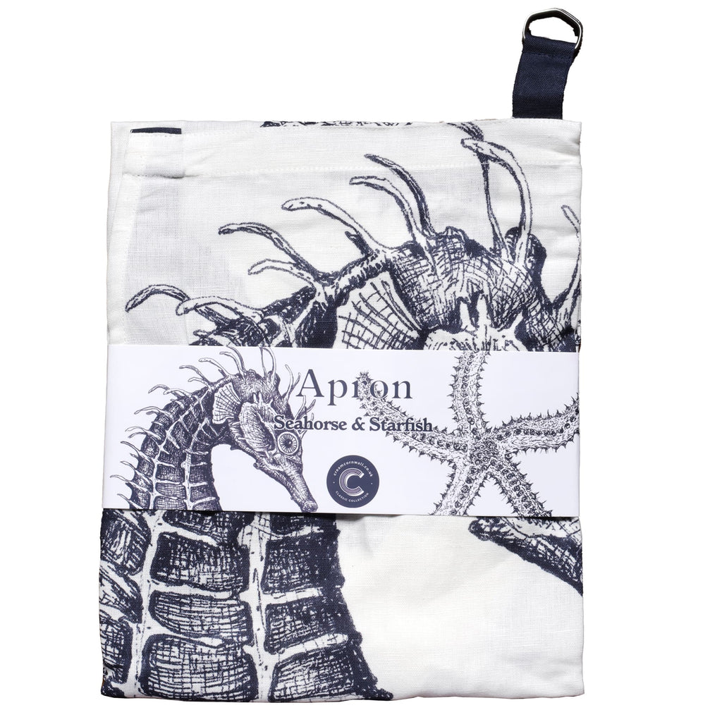 Folded apron with a marine-themed design featuring detailed illustrations of a seahorse and starfish in navy blue on a white background. The apron is wrapped with a branded label that reads 'Apron – Seahorse & Starfish,' showcasing the coastal-inspired artwork.