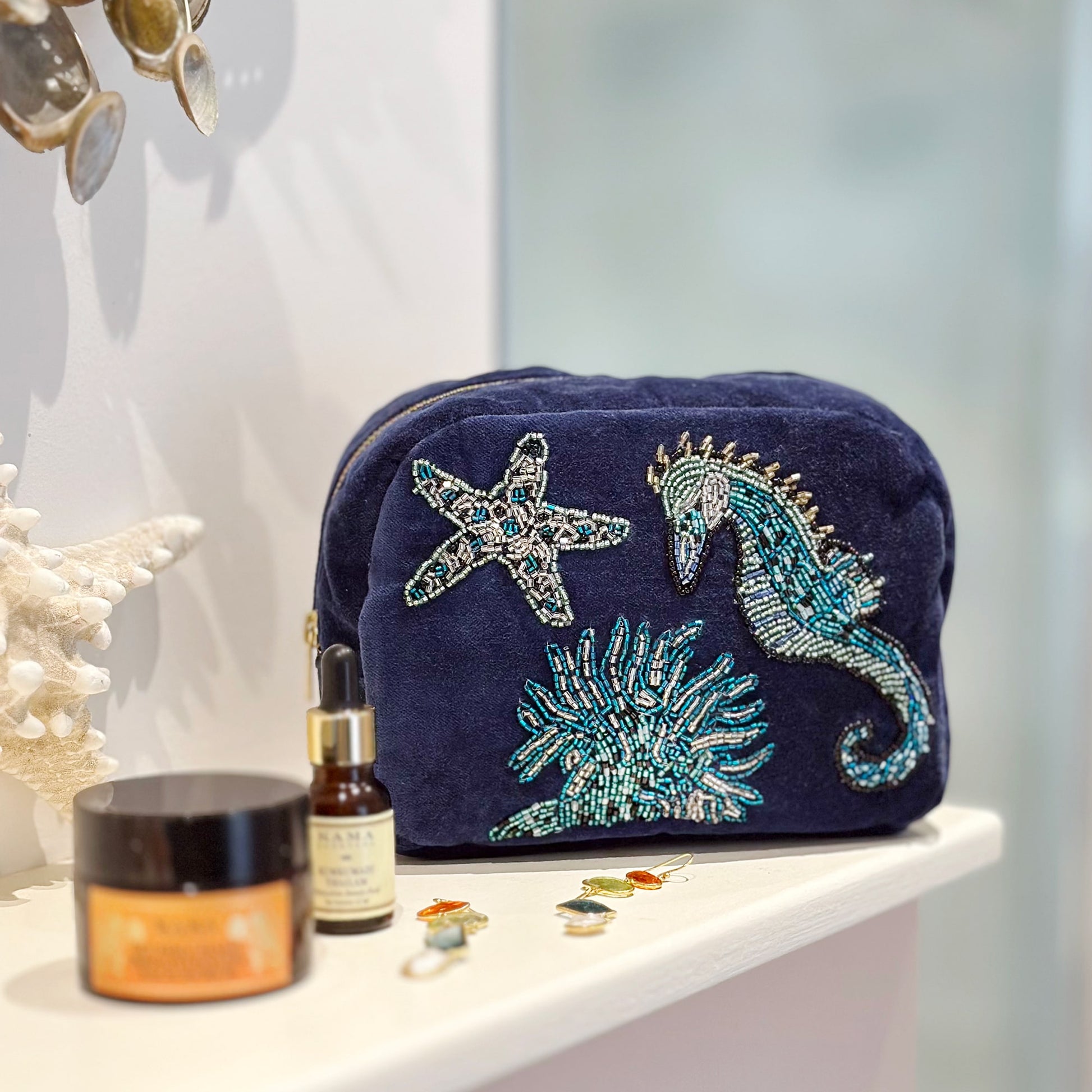 Navy blue velvet cosmetic bag adorned with intricate beaded embroidery of a seahorse, starfish, and coral, displayed on a white bathroom shelf alongside skincare products, seashells, and earrings.