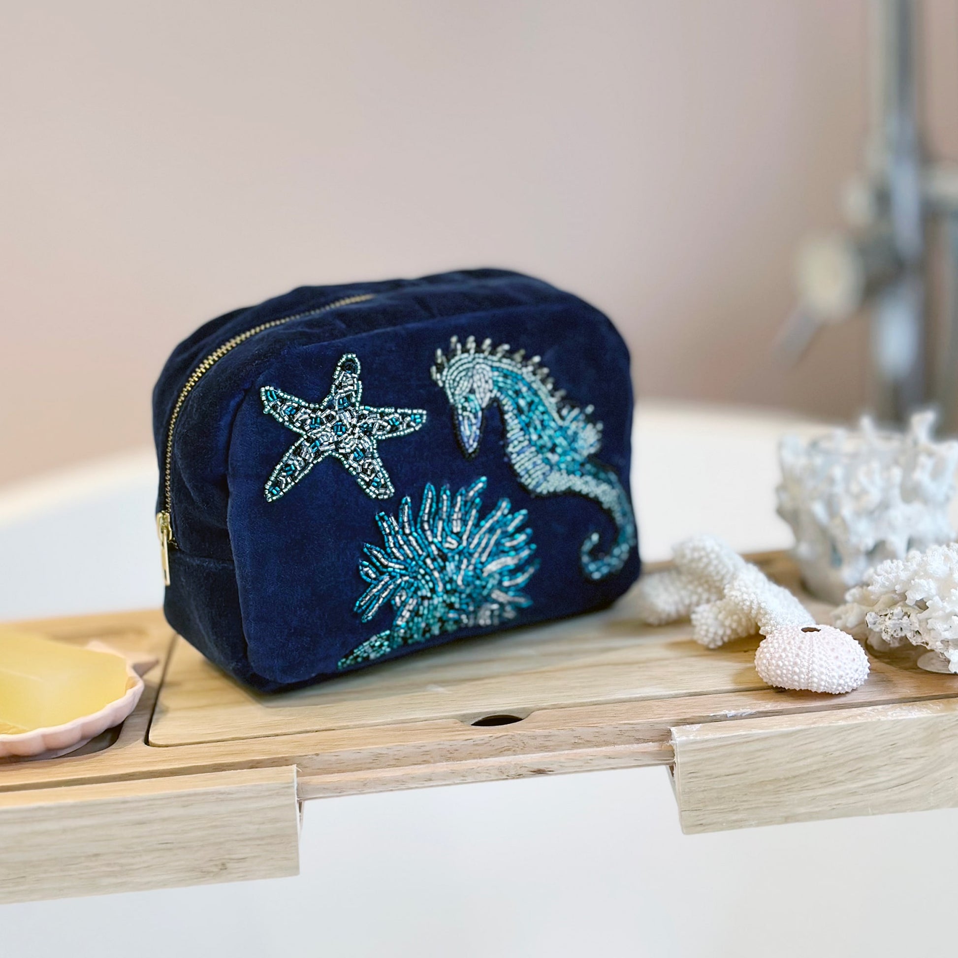 Navy blue velvet cosmetic bag with beaded embroidery of a seahorse, starfish, and coral, placed on a wooden bath tray beside coral decorations and a bar of soap, suggesting a relaxing bathroom setting.