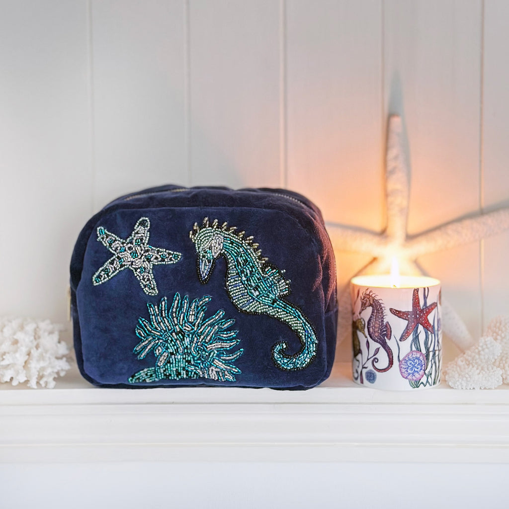 Navy blue velvet cosmetic bag adorned with beaded embroidery of a seahorse, starfish, and coral, displayed on a white shelf beside a lit candle in a matching ocean-themed holder and decorative coral pieces, creating a warm, coastal ambiance.