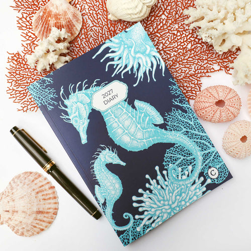 A 2027 daily planner with a dark navy blue cover decorated with light blue seahorse and coral illustrations, styled in a flat-lay with a black pen and seashells on a white background.