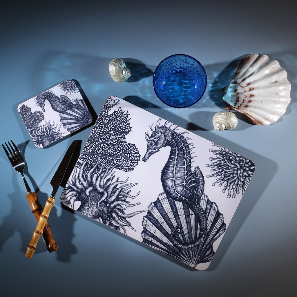 Seahorse Design in Navy on a white Coaster with a matching Placemat.On the table is a bamboo cutlery set and a blue coloured glass