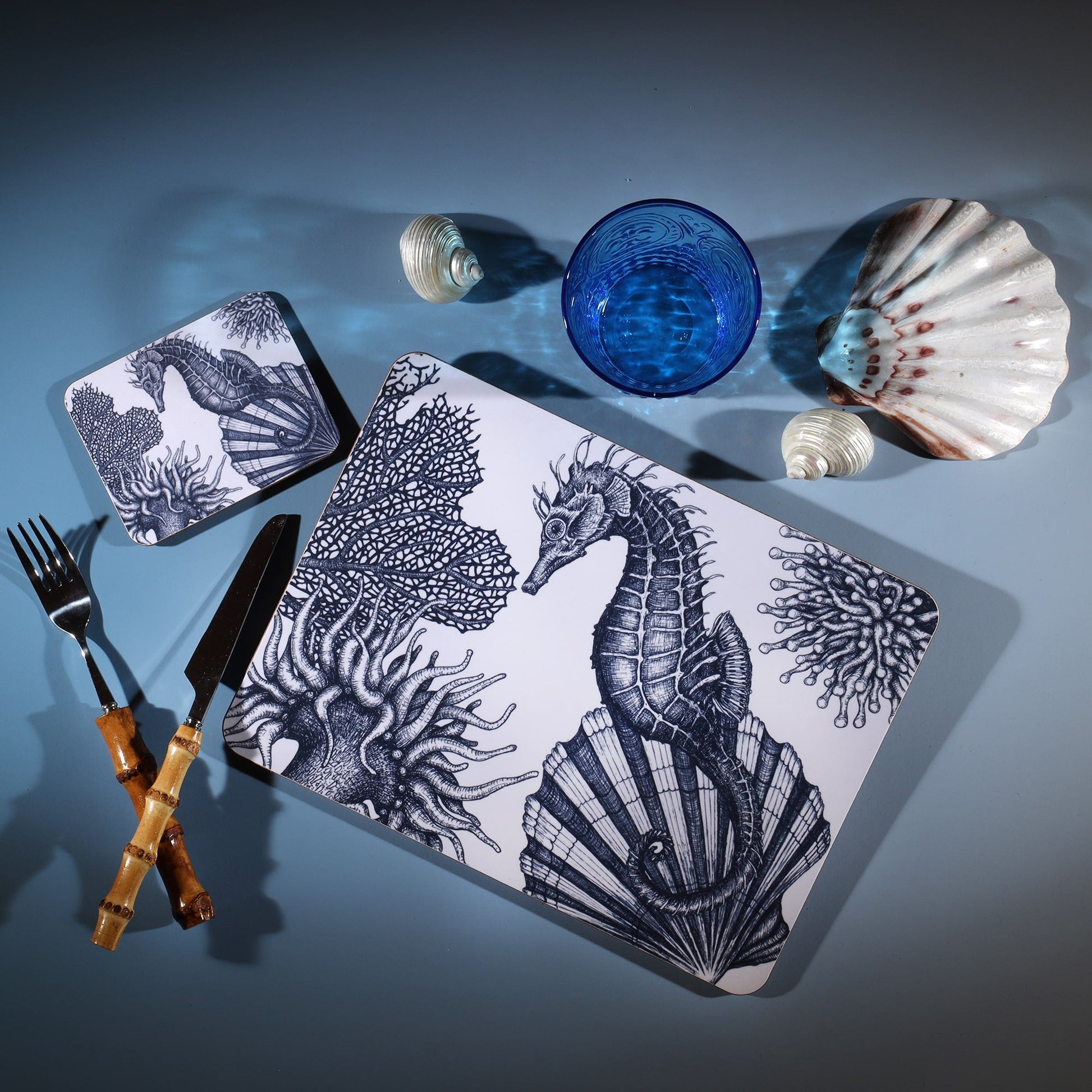 Seahorse Design in Navy on a white Coaster with a matching Placemat.On the table is a bamboo cutlery set and a blue coloured glass