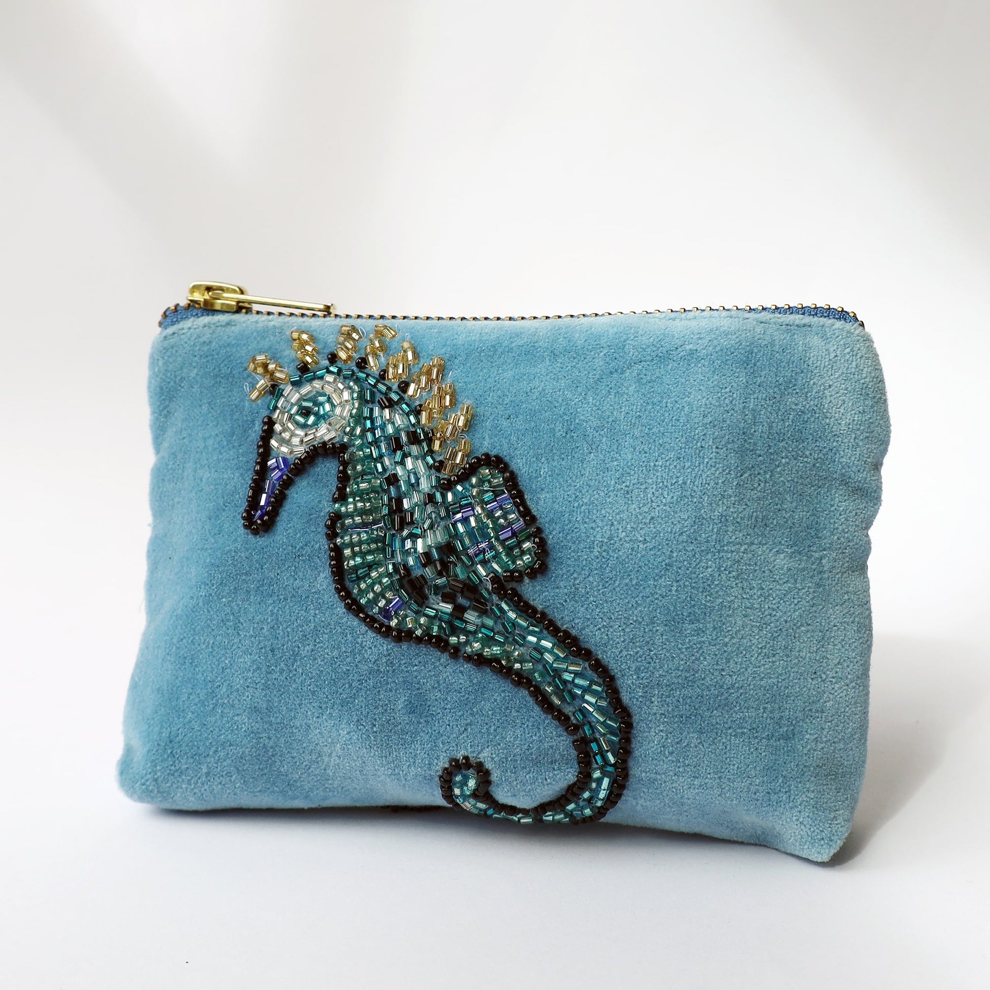 A small light blue velvet purse adorned with a detailed beaded seahorse design. The seahorse is intricately embroidered using beads in shades of teal, turquoise, black, white, and gold, with a curled tail and spiked crest. The purse has a gold zip closure along the top and is photographed against a soft white background, showcasing its luxurious texture and elegant marine-themed embellishment.