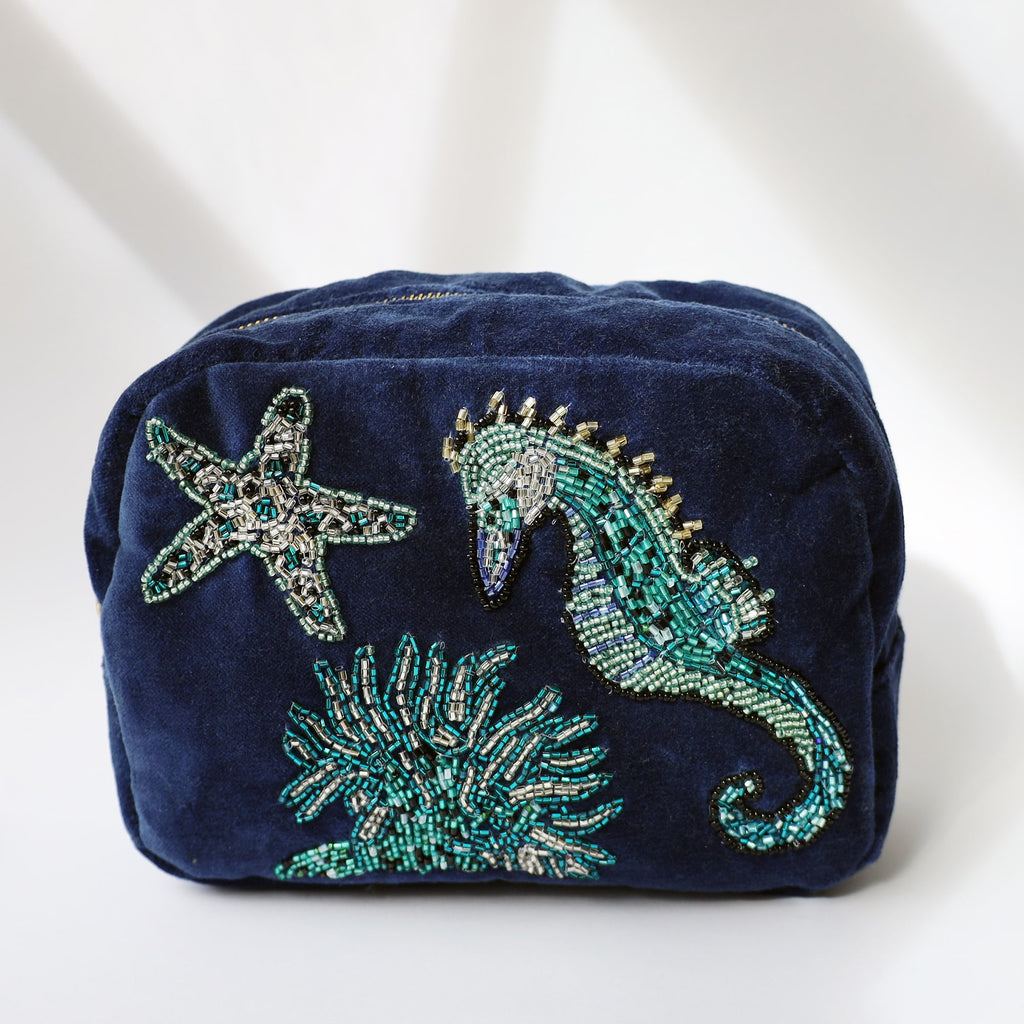 A navy blue velvet cosmetic bag featuring intricate beadwork of an underwater scene, including a seahorse, starfish, and coral. The beaded designs are made with shimmering green, silver, and blue beads, adding texture and a touch of sparkle. The bag is photographed against a soft white background with natural light and subtle shadows.