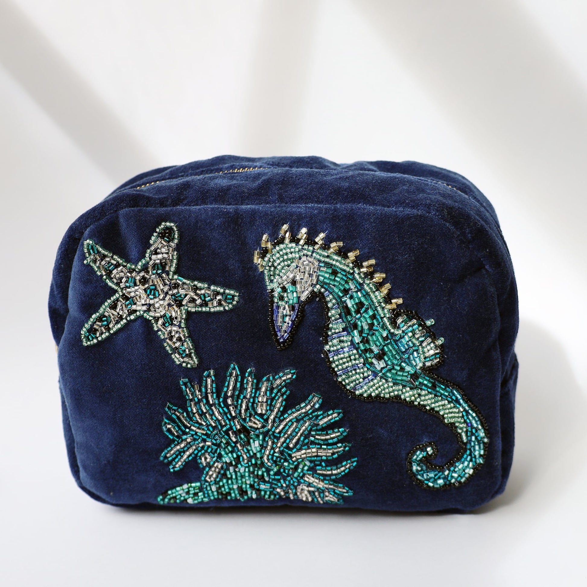 A navy blue velvet cosmetic bag featuring intricate beadwork of an underwater scene, including a seahorse, starfish, and coral. The beaded designs are made with shimmering green, silver, and blue beads, adding texture and a touch of sparkle. The bag is photographed against a soft white background with natural light and subtle shadows.