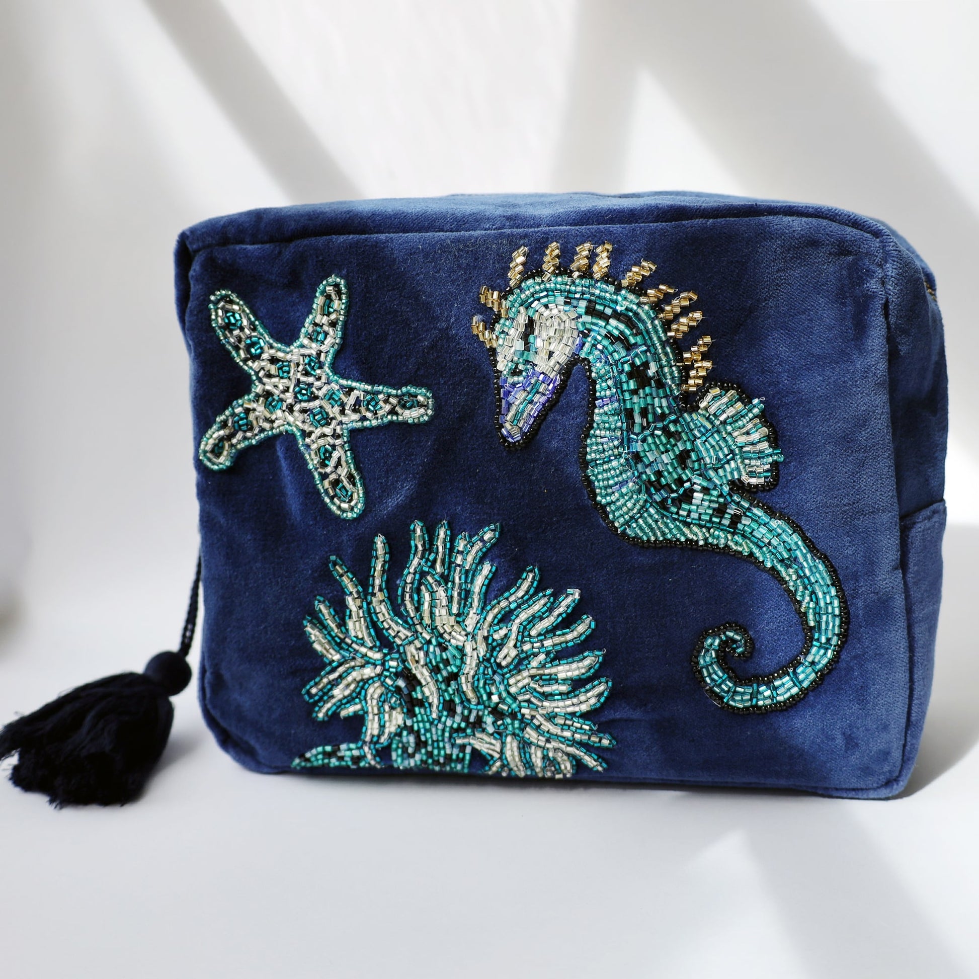 A navy blue velvet washbag featuring intricate beaded embroidery of a seahorse, starfish, and seaweed. The design is composed of shimmering turquoise, silver, gold, and black beads, creating a vibrant underwater scene. The washbag has a black tassel zip pull and is photographed in soft, angled natural light with gentle shadows.