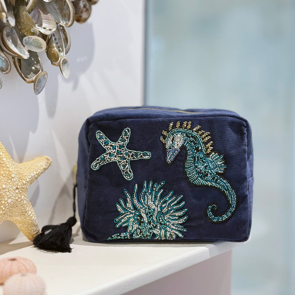 Navy velvet washbag featuring beaded embroidery of a seahorse, starfish, and coral, displayed on a white bathroom shelf with decorative seashells and starfish accents, creating a serene coastal atmosphere.