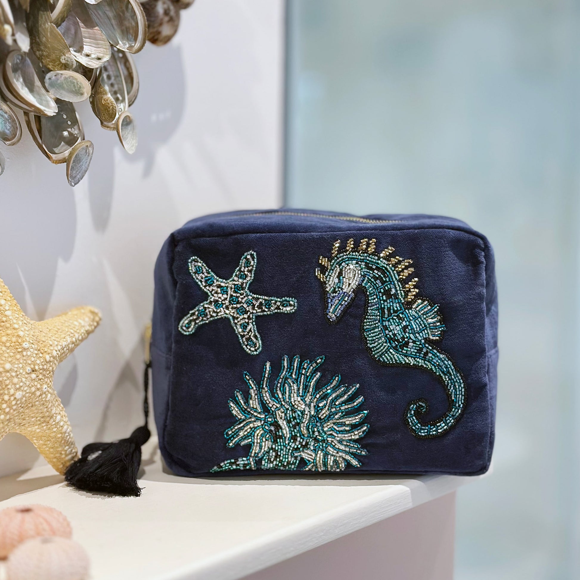 Navy velvet washbag featuring beaded embroidery of a seahorse, starfish, and coral, displayed on a white bathroom shelf with decorative seashells and starfish accents, creating a serene coastal atmosphere.