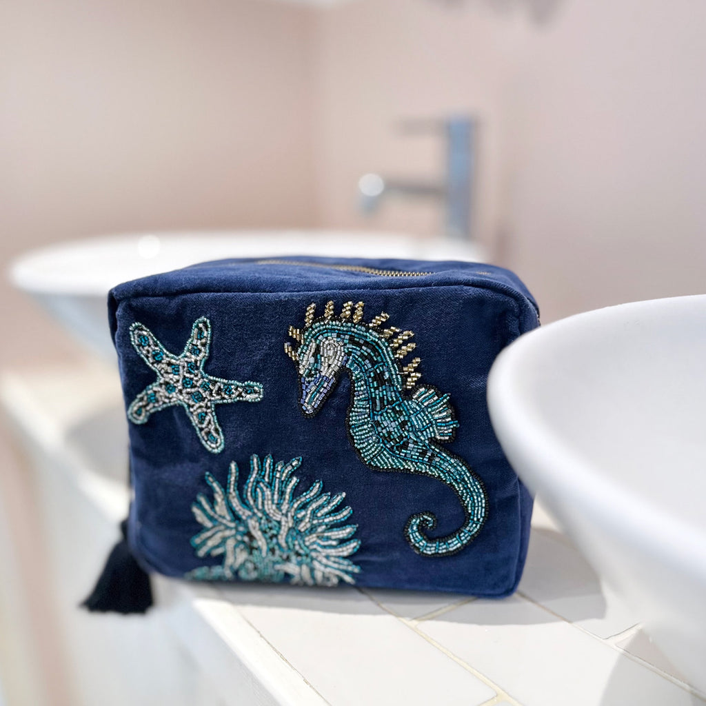 Navy blue velvet washbag adorned with intricate beaded designs of a seahorse, starfish, and coral, placed on a tiled bathroom countertop between two white ceramic sinks, creating a fresh coastal vibe.