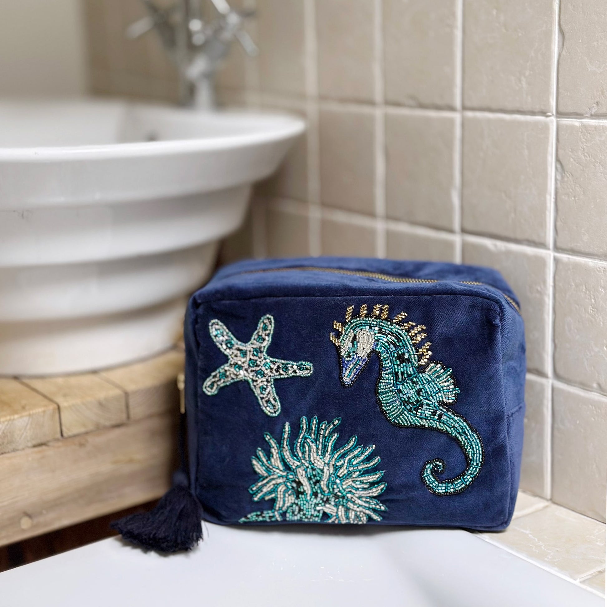 Navy velvet washbag decorated with beaded marine motifs including a seahorse, starfish, and coral, styled in a bathroom next to a ceramic sink and neutral tiled wall, evoking a serene coastal feel.