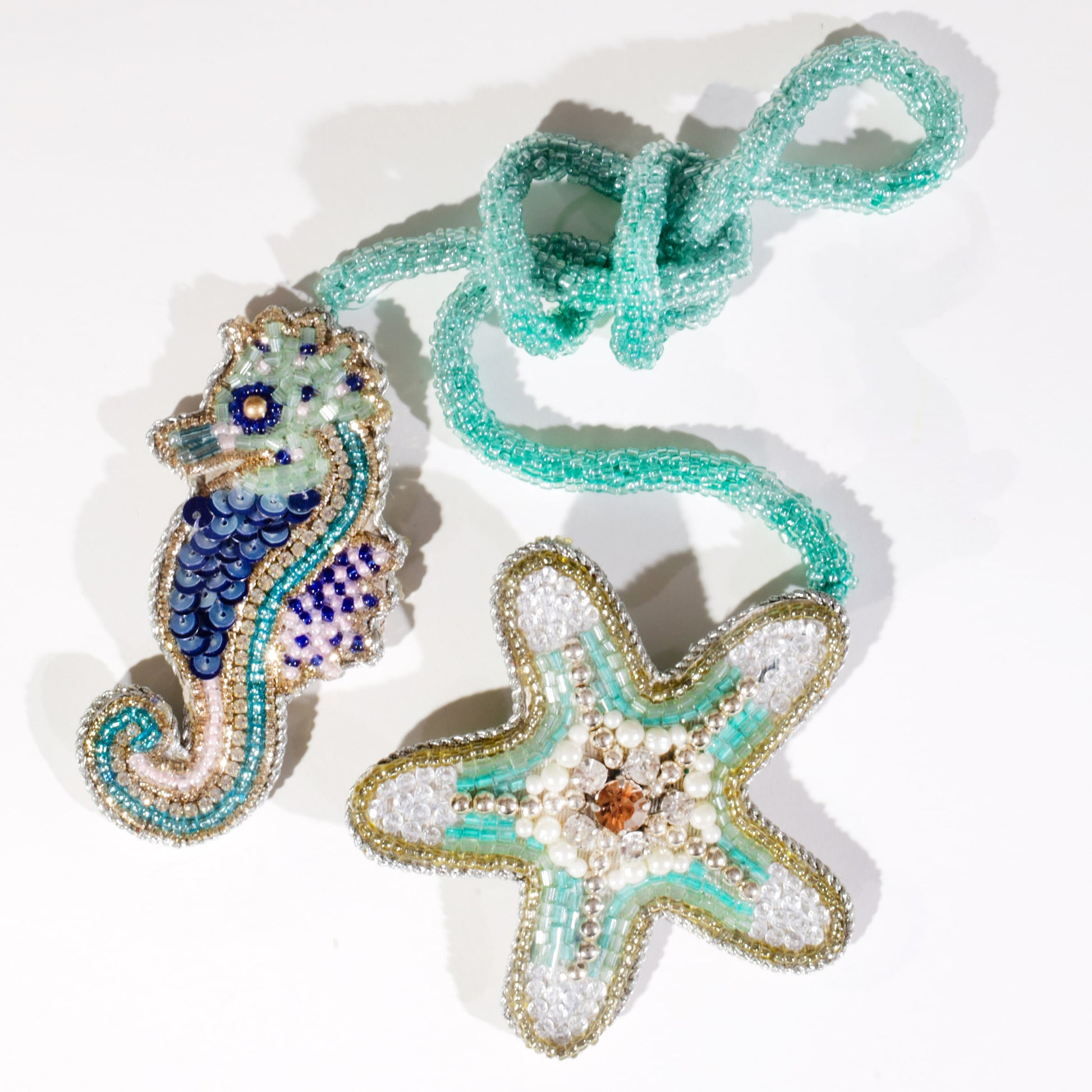 Beaded seahorse and starfish bag charms in shades of turquoise, blue, and gold with pearl and sequin detailing, attached to a looped turquoise beaded cord on a white background.