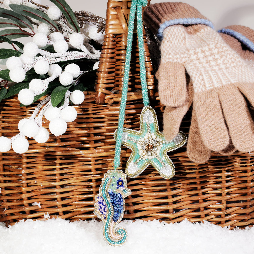 Beaded seahorse and starfish bag charms in turquoise and gold tones hanging from a wicker basket, with a pair of beige winter gloves and frosted white berries, set on faux snow.