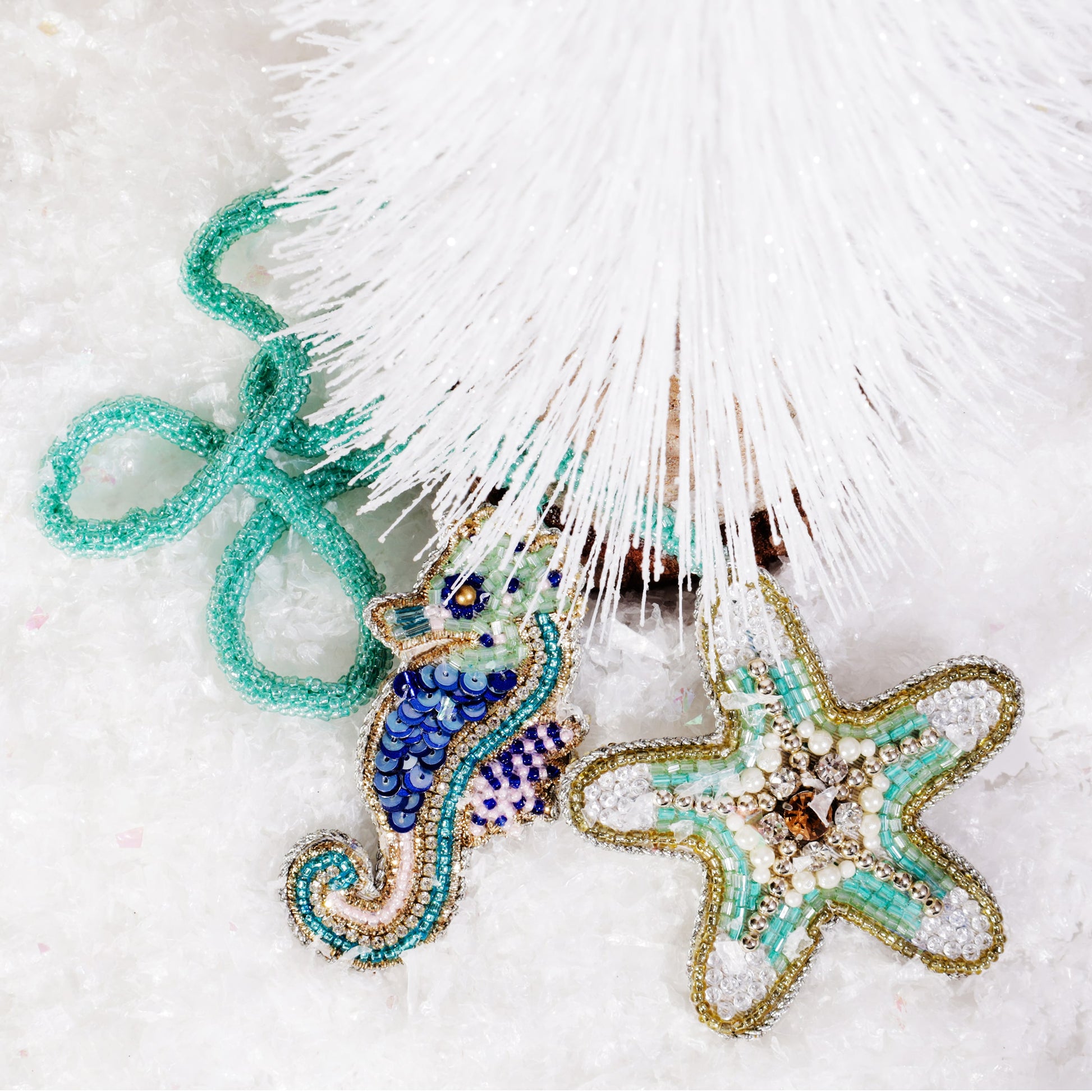 Beaded seahorse and starfish bag charms in shades of turquoise, blue, and gold resting beneath a white frosted Christmas tree, surrounded by sparkling artificial snow.