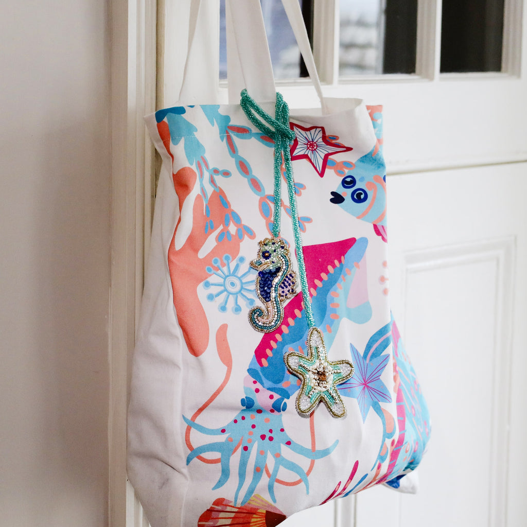 Colourful ocean-themed tote bag with coral and sea creature patterns, decorated with beaded seahorse and starfish bag charms in turquoise, blue, and gold tones hanging from the handle.