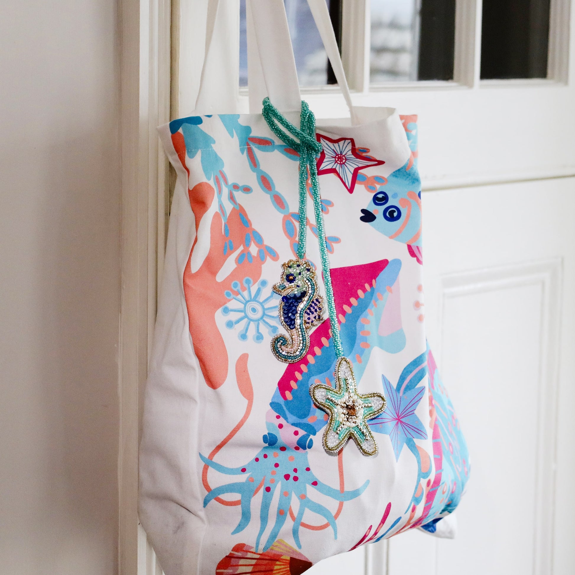 Colourful ocean-themed tote bag with coral and sea creature patterns, decorated with beaded seahorse and starfish bag charms in turquoise, blue, and gold tones hanging from the handle.