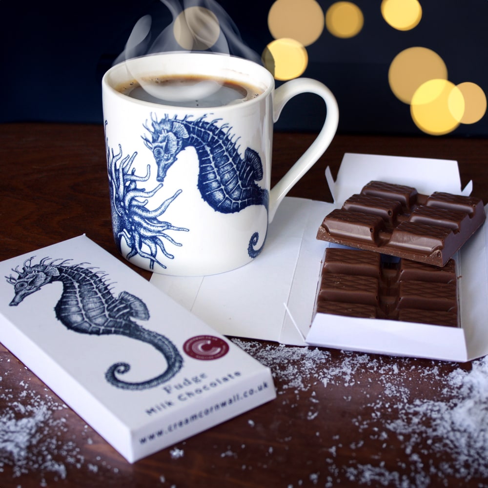 Alt text: White ceramic mug featuring a blue seahorse and anemone design, filled with steaming coffee, placed beside a partially unwrapped bar of milk chocolate on a wooden surface dusted with faux snow and warm festive lights in the background.