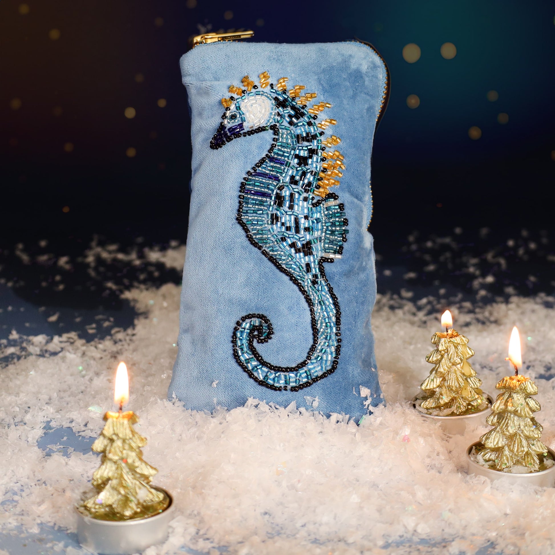 Light blue velvet glasses case featuring an intricate beaded seahorse design, surrounded by faux snow and small golden Christmas tree candles with a softly glowing festive background.