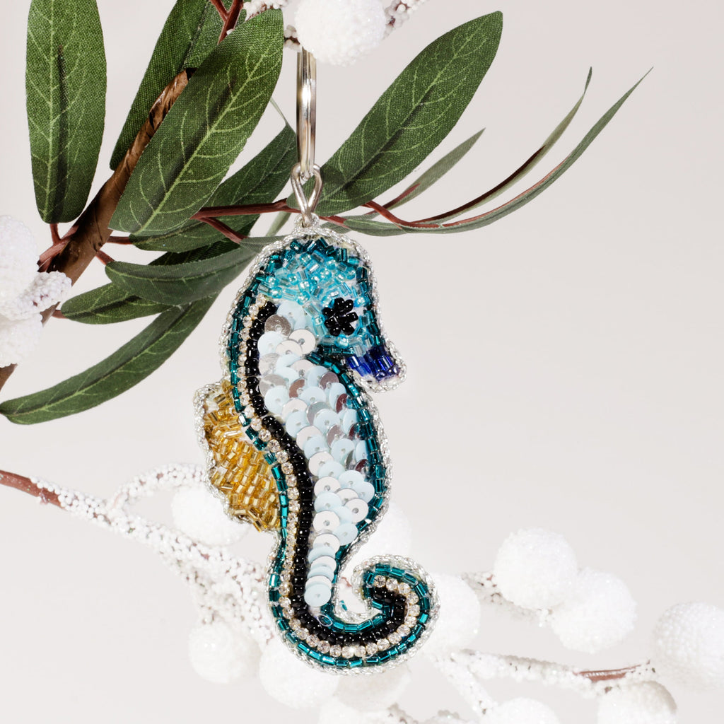 A beaded seahorse keyring with turquoise, silver, black, and gold sequins hanging from a branch with green leaves and white frosted berries against a light background.